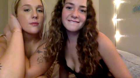 Media: Video of two young women with fair skin and long hair, one with blonde and the other with curly brown locks. They wear black lingerie, smiling, in a softly lit room with string lights.