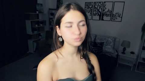 Media: Video of a young woman with long brown hair and fair skin, wearing a strapless green top, blowing a kiss in a dimly lit room with a black tree art print on the wall.