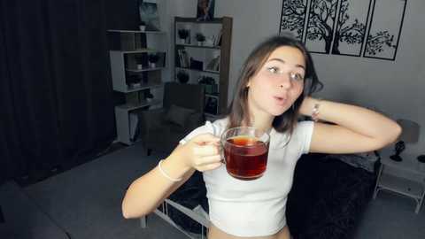 Media: Video of a young woman with light skin, brown hair, and a white crop top, drinking tea in a dimly lit, minimalist bedroom with dark curtains, black bedspread, and white shelves.