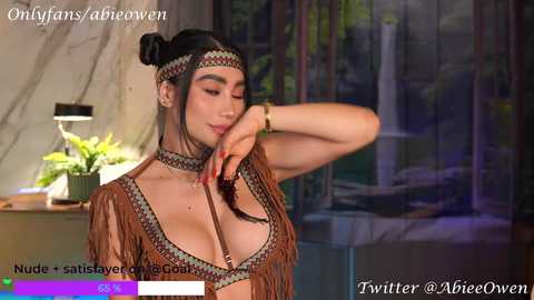 Media: Video of a woman with medium skin tone, dark hair in a bun, wearing a tribal-inspired outfit with fringe and a headband. She poses seductively in front of a jungle mural.