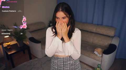 Media: Video of a young woman with long black hair, wearing a white top and patterned skirt, covering her face in a living room with beige furniture and a floral arrangement.