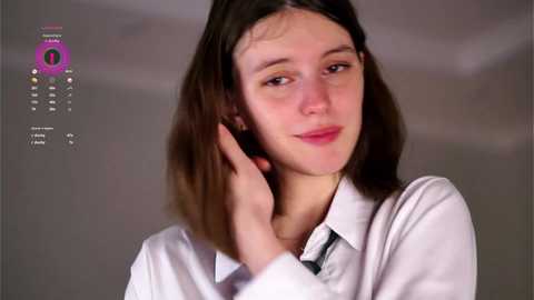 Media: Video of a young woman with straight brown hair, wearing a white shirt, smiling slightly, with a blurred background.