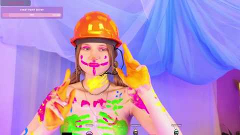 Media: Video of a young woman with fair skin and brown hair, wearing an orange helmet, covered in vibrant paint. She poses playfully, with a smile, surrounded by pastel-colored drapes and soft lighting.