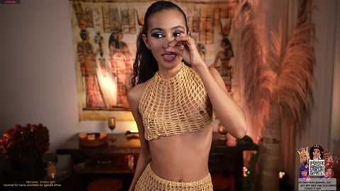 Media: Video of a young woman with medium-brown skin and a slim, toned physique, wearing a gold crochet halter top and matching high-waisted shorts. She poses in a cozy, dimly lit room adorned with African art and candles.