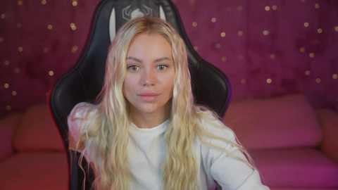 Media: Video of a young woman with long, wavy blonde hair, light skin, and blue eyes, seated in a black gaming chair, wearing a white shirt. Background features a plush, pink velvet tufted headrest and wall adorned with golden glitter.