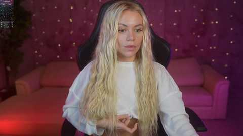 Media: Video of a young woman with long, wavy blonde hair, fair skin, and light makeup, sitting in a black gaming chair, wearing a white long-sleeve top. Background features a pink tufted sofa and wall with small, glowing lights.