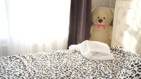 Media: Video of a bedroom with a large, fluffy white teddy bear with a red bow on a leopard-print bedspread, against white curtains and a tufted headboard.