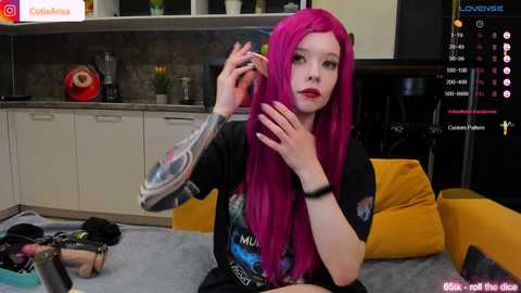 Media: Video of a pale-skinned woman with vivid pink hair, wearing a black \"Mushroom\" t-shirt, holding a cigarette, in a modern kitchen with yellow cushions.