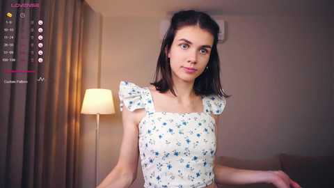 Media: A video of a young woman with fair skin, brown hair, and blue eyes, wearing a white floral dress, standing indoors next to a lamp and beige curtains.