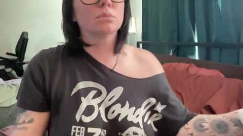 Media: Video of a fair-skinned woman with dark hair, glasses, wearing a dark grey off-shoulder \"Blondie\" T-shirt, sitting on a bed with red and green bedding.