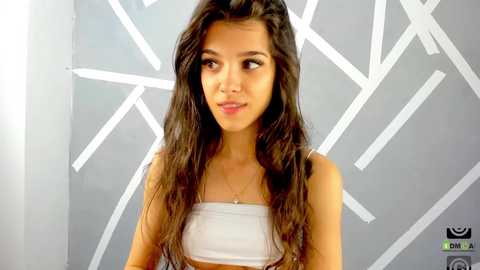 Media: Video of a young woman with long, wavy brown hair, wearing a white bandeau top, standing against a geometric, white-painted wall. She has a light skin tone and is smiling slightly.