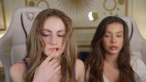 Media: Video of two young women in a well-lit room, one with long blonde hair and a contemplative expression, the other with long dark hair and a neutral expression.