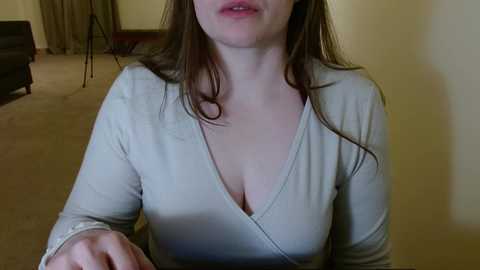 Media: A video shows a woman with long brown hair, wearing a low-cut beige top, sitting on a beige carpeted floor. Her face is partially visible, and a hand is extended towards the camera. The background features a dark couch and beige walls.