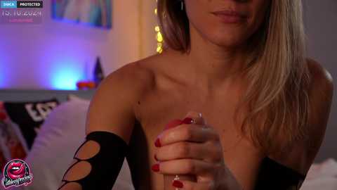 Media: Video of a topless blonde woman with fair skin, medium breasts, and red-painted nails, holding a lit cigarette in a dimly lit bedroom.