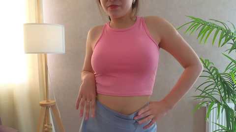 Media: Video of a slim, light-skinned woman with medium-sized breasts in a pink sleeveless crop top and light blue pants, standing in a softly lit room with a potted plant and a floor lamp.