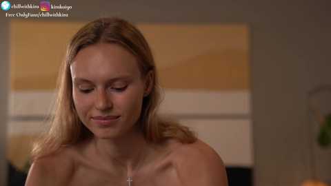 Media: A video of a young, fair-skinned, blonde woman with a slim figure, topless, gazing downwards in a softly lit room with a yellow and white abstract wall painting.