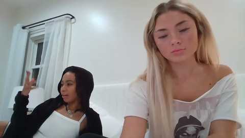 Media: A video of two young women in a bedroom. One is a dark-skinned woman with a hijab, black jacket, and white top, gesturing with her right hand. The other is a blonde woman in a white graphic tee, off-shoulder, looking down.