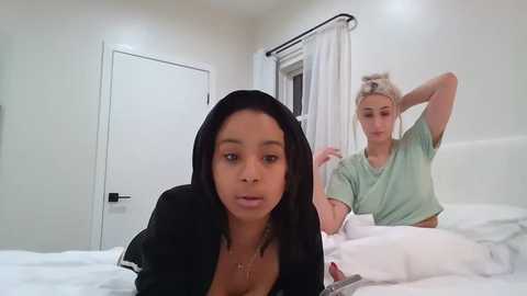 Media: A candid photo of a dark-skinned woman with straightened black hair, wearing a black robe, lying on a white bed in a white bedroom, with a blonde woman in a light green shirt in the background.