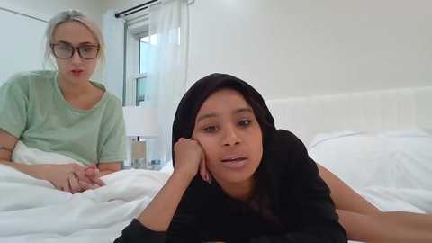 Media: Video of a woman with short blonde hair and glasses, wearing a light green t-shirt, sitting on a bed in a hospital room. Beside her, a young woman with dark skin, wearing a black hijab and black top, lies on her stomach, looking sad.