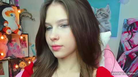 Media: Video of a young woman with long brown hair and fair skin, wearing a pink shirt, in a room with colorful posters, stuffed animals, and string lights.