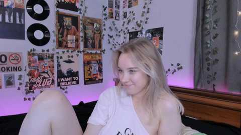 Media: A video of a smiling, fair-skinned, blonde woman in a white off-shoulder T-shirt, seated on a bed with posters and records on the wall.