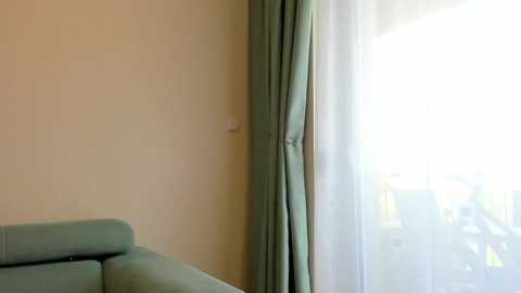 Media: Video of a corner of a minimalist room with a light green sofa and matching curtains, next to a sheer white curtain partially covering a window that reveals a bright, outdoor scene.