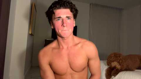 Media: Video of shirtless, muscular, tanned white man with curly brown hair, slightly red face, sitting in a dimly lit bedroom with a brown dog on a bed.