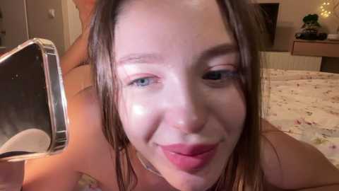 Media: A close-up video of a young woman with light skin and blue eyes, smiling, in a dimly lit bedroom. She has wet hair and is holding a phone.