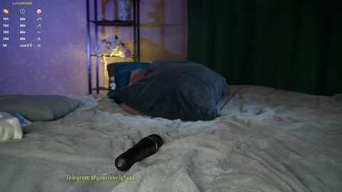 Media: Video of a dimly lit bedroom with a person curled up under a gray blanket on a bed, a black remote on the bed, and a green curtain in the background.