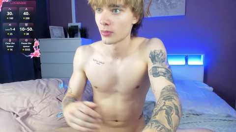 Media: Video of a shirtless, slender, young man with light skin, blond hair, and tattoos on his arms, sitting on a bed with a pink blanket in a dimly lit bedroom.