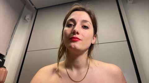 Media: Video of a young Caucasian woman with fair skin, brown hair, wearing a gold necklace, standing topless in a bathroom with white tiled walls.