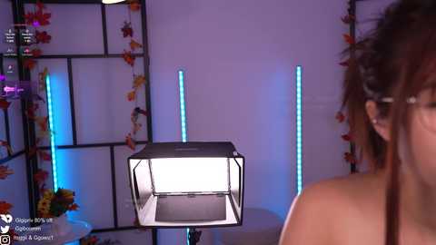 Media: Video of a woman with dark hair, wearing glasses, standing in a modern room with a black-framed lightbox and colorful wall decorations.