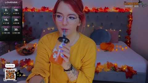 Media: A young woman with fair skin and red hair, wearing glasses, sips an orange beverage, surrounded by Halloween decorations on a bed.