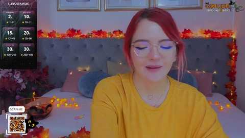 Media: Video of a fair-skinned, red-haired woman with glasses in a yellow shirt, sitting on a bed decorated with fall-themed candles, flowers, and a \"LOVESE\" watermark.