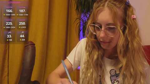 Media: Video of a young, blonde woman with wavy hair, wearing glasses and a white T-shirt, seated at a desk, with a computer screen displaying social media metrics in the background.