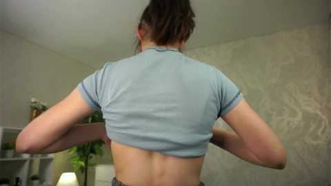 Media: Video of a woman with fair skin, brown hair tied back, lifting her light blue cropped top, revealing her back and toned physique. Background features a light gray textured wall and a white shelf with green plants.