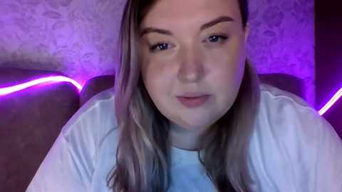 Media: Video of a young Caucasian woman with light skin, shoulder-length wavy blonde hair, and light makeup. She wears a white shirt, sitting on a dark brown couch with purple LED strip lights in the background.