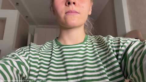 Media: Video of a young woman with fair skin, wearing a green and white striped shirt, captured from a low angle, showing her upper body and face. Background features a modern, beige-toned living room with white walls and recessed lighting.