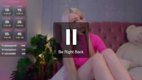 Media: Video of a blonde woman in a pink sweater, legs crossed, sitting on a bed with a teddy bear, indoor background with a digital clock and plant.