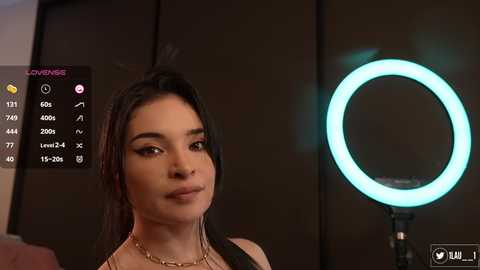 Media: Video of a young woman with long black hair, fair skin, wearing a black top and necklace, smiling. Background shows a glowing ring light and digital camera settings.