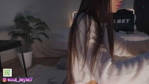 Media: Video of a woman with long, dark hair, sitting on a bed with a white sheet, wearing a white sweater, in a dimly lit room with a potted fern, a black bag, and a \"BET\" pillow visible.