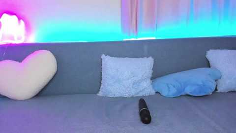 Media: A video of a gray sofa with three pillows (white, blue, and fluffy white) and a black dildo on the floor, under soft, colorful LED strip lighting.