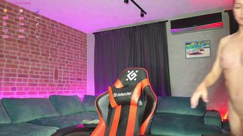 Media: Video of a modern living room with a brick wall, purple lighting, a green couch, and a gaming chair. A nude person, likely male, walks in the background.