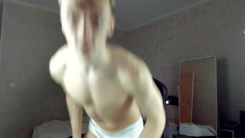 Media: Video of a shirtless, muscular man with light skin and blonde hair, wearing white underwear, captured from behind in a dimly lit bedroom with beige walls and a large mirror.