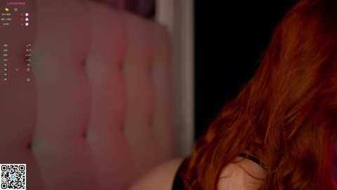 Media: A video of a woman with long, wavy red hair, wearing a black top, seen from the side, in a dimly lit room with a blurred, pinkish, textured wall in the background.