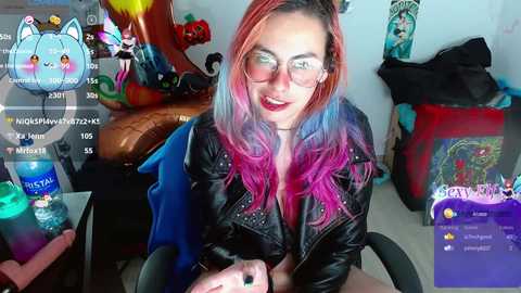 Media: Video of a light-skinned woman with long, pink and blue hair, wearing glasses and a black leather jacket, sitting in a cluttered room with colorful decorations and gaming setup.