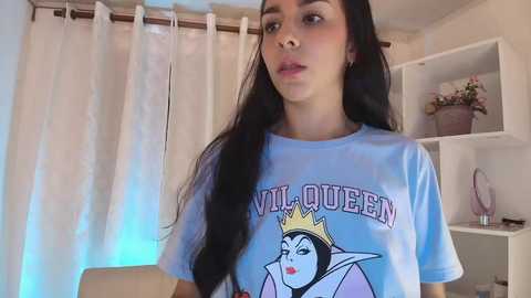 Media: A young woman with long, straight black hair, fair skin, and dark eyes, wearing a light blue \"Villain Queen\" t-shirt featuring a cartoon image of Maleficent. Background includes white shelves and a floral arrangement.