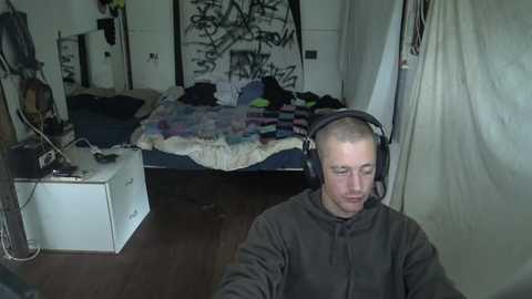 Media: Video of a young, bald man in a gray hoodie, wearing headphones, sitting on a wooden floor in a messy, graffiti-covered room with a bed and various items.