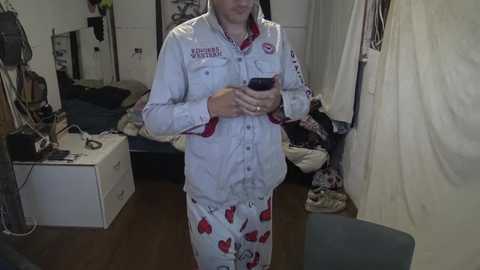 Media: Video of a person in white pajamas with red heart patterns, holding a smartphone, standing in a cluttered bedroom with a bed, dresser, and clothes.