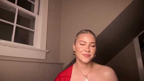 Media: Video of a smiling, light-skinned woman with blonde hair tied back, wearing a red off-shoulder dress, gold earrings, and a gold necklace. She stands in a beige-walled room with a white-framed window and dark wooden staircase in the background.